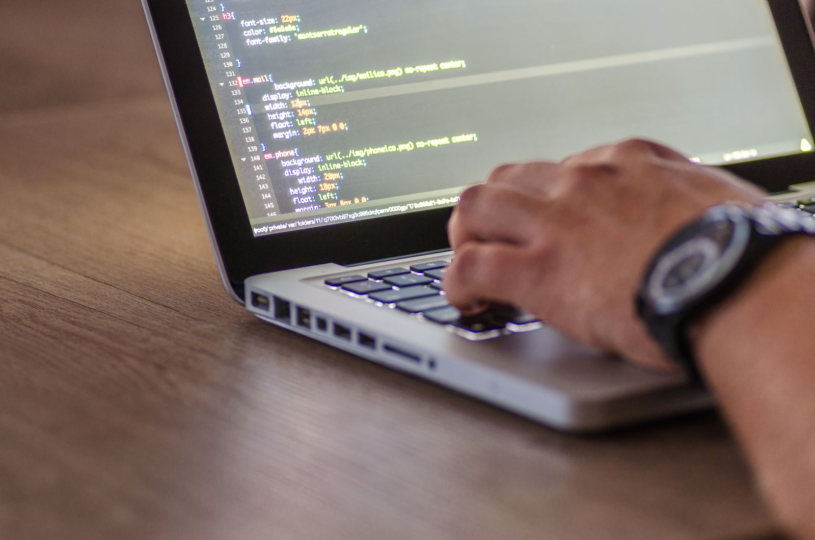 Uma mão digita no teclado de um notebook sobre uma mesa de madeira enquanto a tela mostra linhas de código.