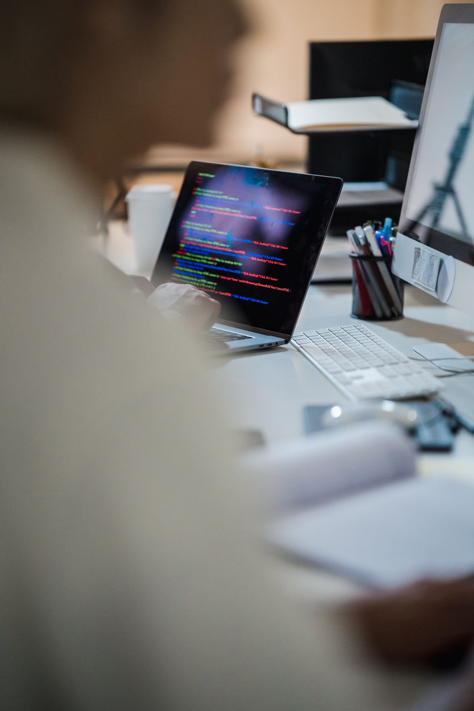 Pessoa desfocada trabalha em um notebook com código na tela, em um escritório com monitor e porta-canetas.