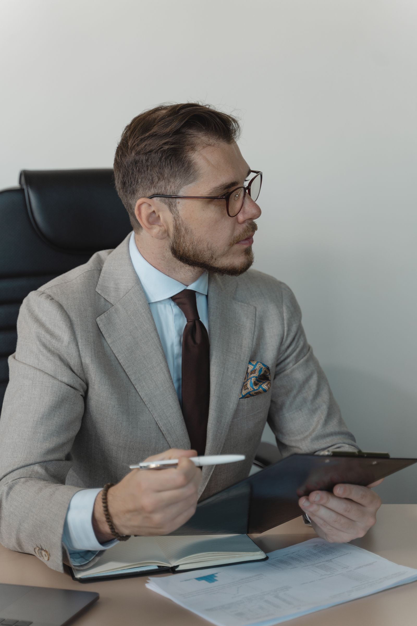 Homem de barba, terno cinza e óculos sentado a uma mesa, segurando caneta e prancheta e olhando para o lado.