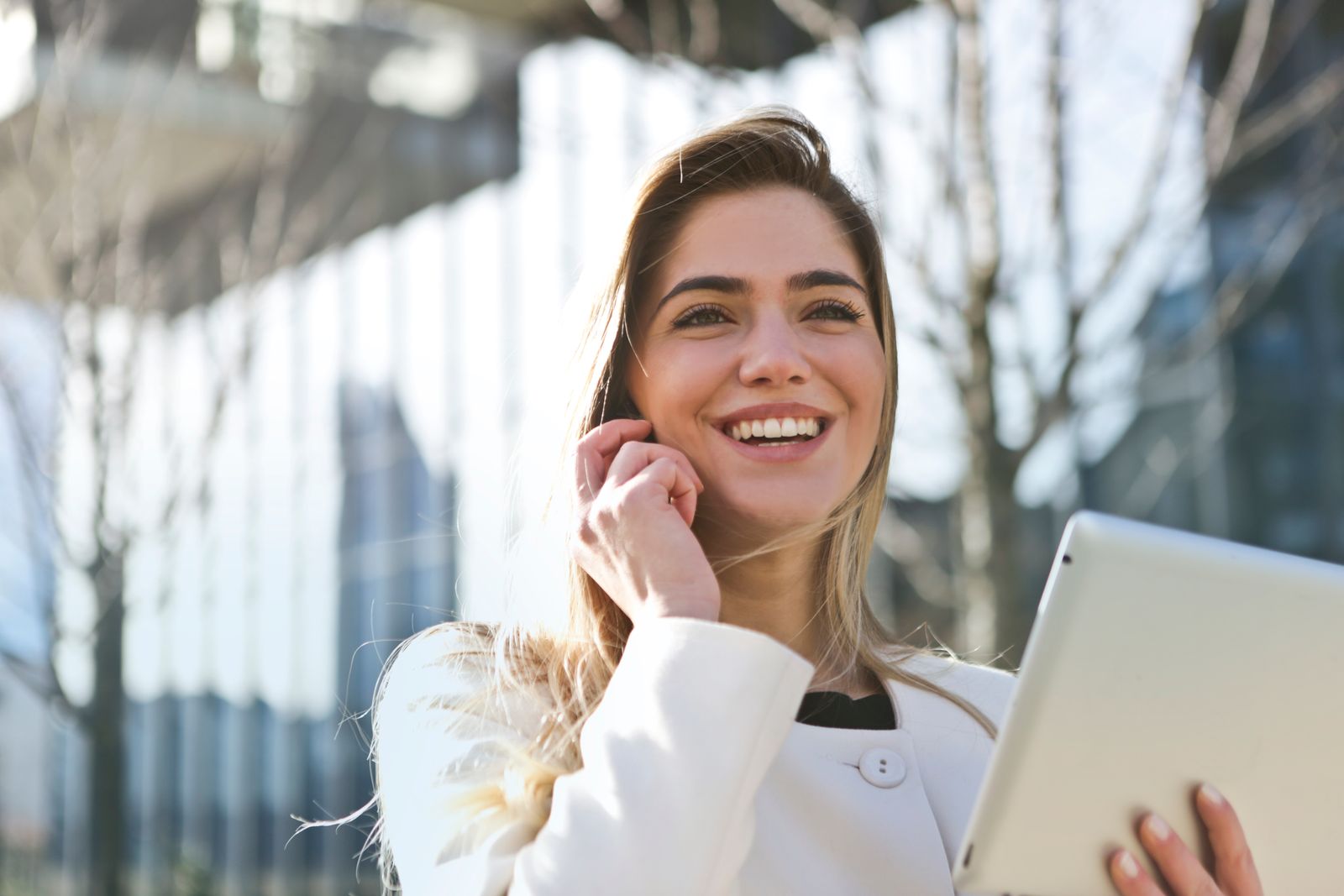 Mulher sorridente de casaco branco fala ao telefone e segura um tablet ao ar livre, diante de prédios de vidro.